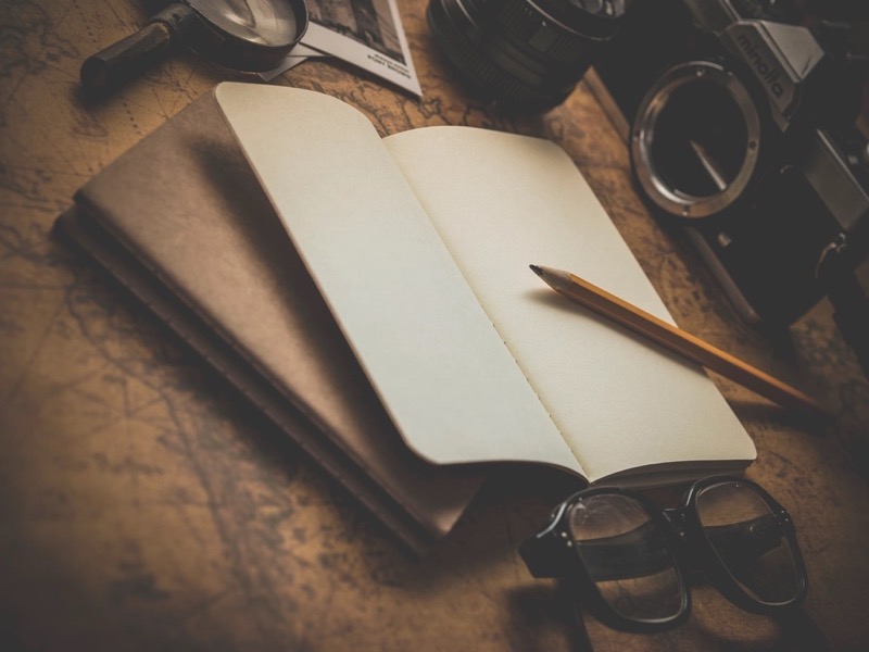 An open notebook and pencil resting on an old map beside a film camera and glasses